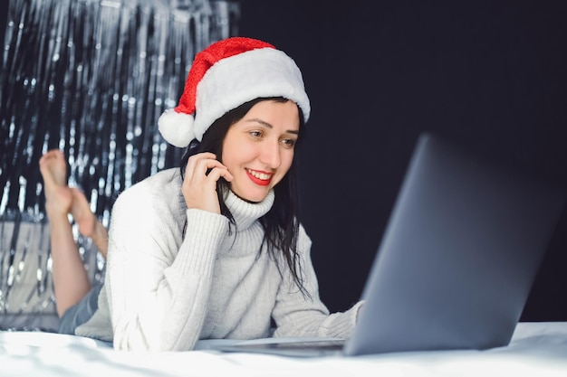 Belle femme brune souriante excitée en pyjama rouge se trouve dans son lit en chapeau de père noël du nouvel an