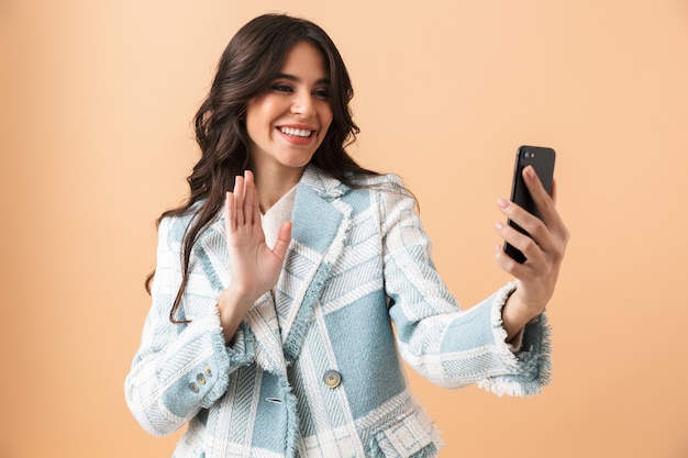 Belle femme brune vêtue d'une veste à carreaux debout isolé sur beige, prenant un selfie avec un téléphone mobile