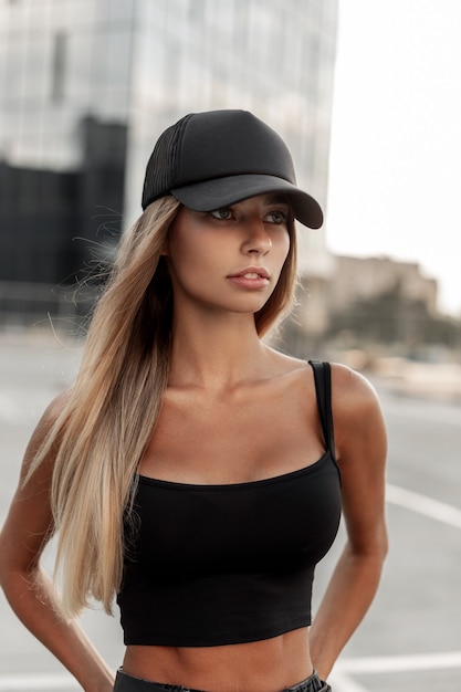 Photo belle femme butin avec une casquette noire à la mode dans un t-shirt noir marche dans la rue près des bâtiments modernes