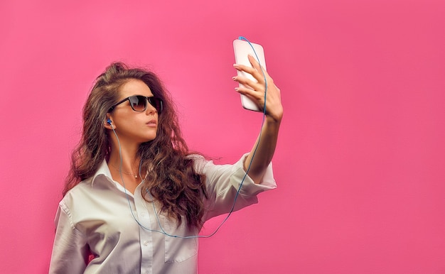 Belle femme caucasienne dans une chemise blanche dans des verres avec des écouteurs, tenant une tablette blanche dans les mains et fait selfie photo sur un mur lumineux rose.