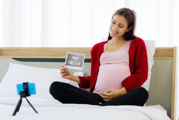 Photo belle femme caucasienne enceinte montre avec bonheur une image échographique lors d'un appel vidéo via un smartphone