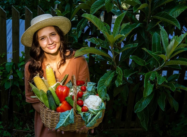 Belle femme caucasienne portant un chapeau de paille et une robe de lin dans le jardin avec un panier plein de légumes