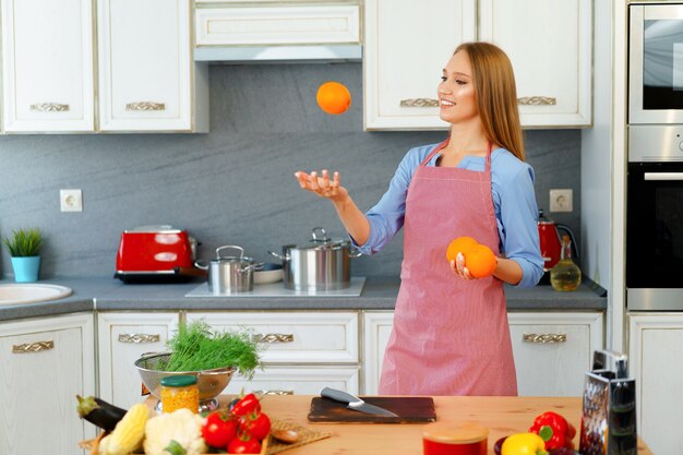 Belle femme caucasienne en tablier rouge tenant des oranges mûres en se tenant debout dans la cuisine