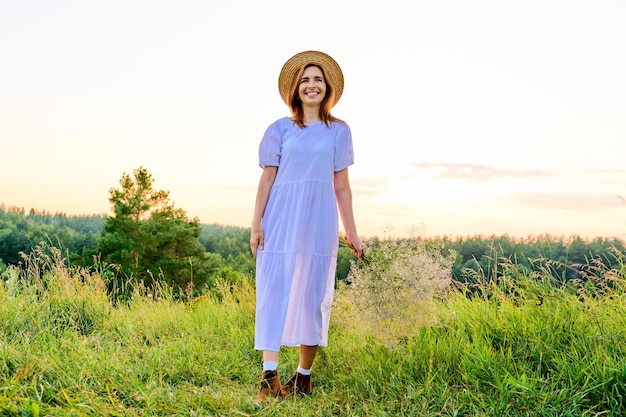 Belle femme en chapeau de robe blanche avec bouquet de fleurs sur le pré au coucher du soleil