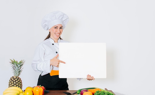 Belle femme chef avec table de légumes tenant un tableau blanc Chef femme avec des légumes à table tenant un menu vierge Fille chef dans la cuisine montrant un tableau blanc