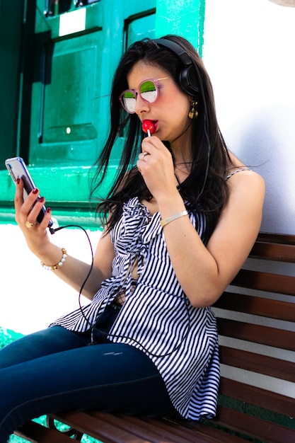 belle femme colombienne porte des lunettes de soleil et écoute de la musique de son téléphone portable avec des écouteurs assis sur une chaise en bois mangeant un bonbon rouge