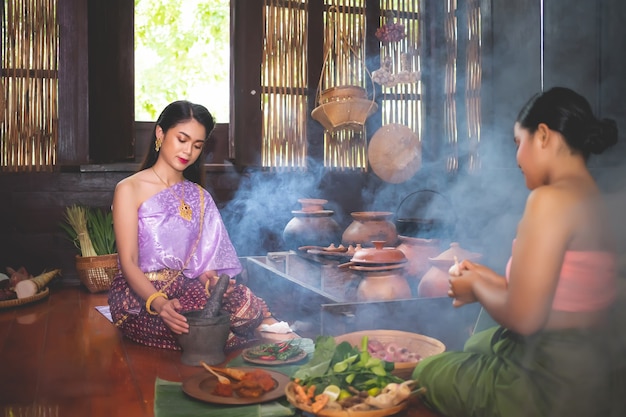 Une belle femme en costume traditionnel thaïlandais utilise un mortier pour piler la pâte de piment dans la cuisine avec un serviteur assis à côté de la préparation des ingrédients. Dans le concept de la cuisine des Thaïlandais