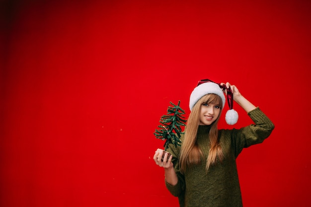 Une belle femme dans un chapeau de santa tient un petit arbre de Noël dans ses mains sur un fond rouge