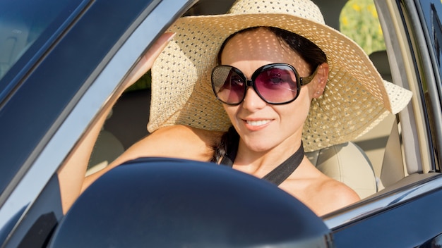 Belle femme dans un chapeau de soleil de paille à larges bords et lunettes de soleil assis dans le siège du conducteur d'une voiture à moteur