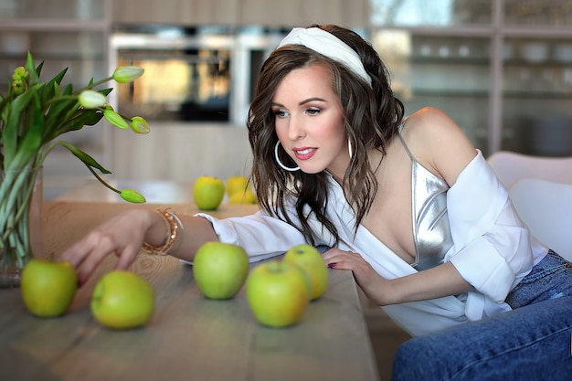 Belle femme dans une chemise blanche et un jean bleu à une table avec des pommes vertes. Portrait d'une jeune femme dans un intérieur blanc.