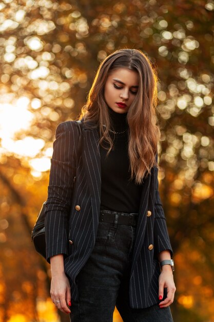 Belle femme dans un costume élégant à la mode avec une veste noire, un pull et un jean avec un sac à dos se promène dans un parc d'automne au coucher du soleil