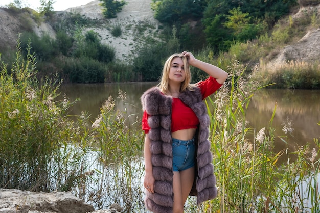 Belle femme dans un manteau de fourrure et un T-shirt et un short rouge clair posant au bord du lac par une chaude journée d'automne