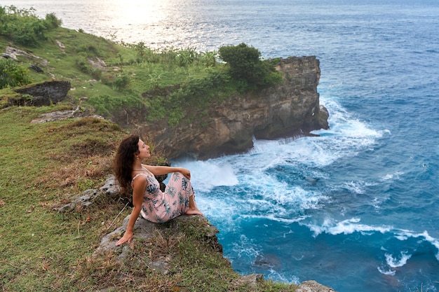 Photo une belle femme dans une robe rose est assise sur une falaise au-dessus de l'océan sur l'île de nusa penida devils billabong un lagon incroyablement merveilleux avec des éclaboussures des vagues