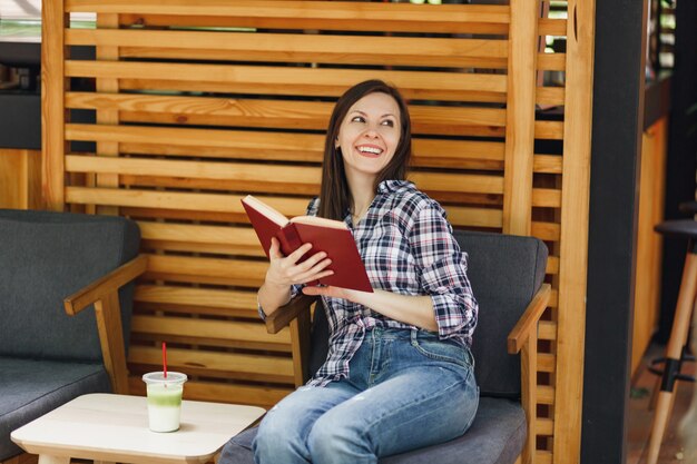 Belle femme dans la rue en plein air café d'été café en bois assis dans des vêtements décontractés, livre de lecture avec une tasse de cocktail