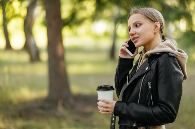 Une belle femme dans une veste en cuir se promène dans le parc et parle au téléphone avec un verre de café à la main