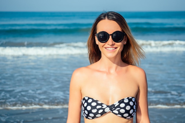 Belle femme debout en maillot de bain et portant des lunettes de soleil sur la plage.