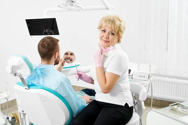 Belle femme dentiste tenant un miroir pour un client masculin d'une dentisterie qui sourit en vérifiant ses dents.