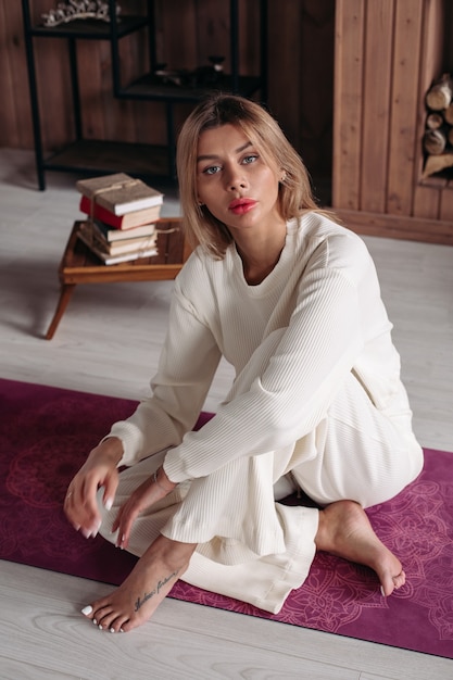 Une belle femme détendue aux cheveux blonds en vêtements blancs est assise sur le tapis de yoga et regarde de côté