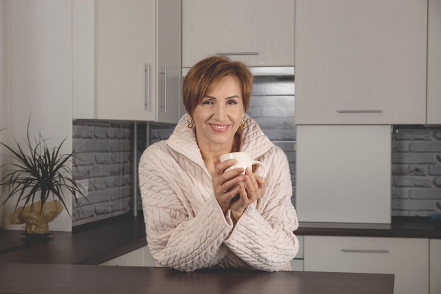 Belle femme du baby-boom d'âge moyen tenant une tasse et un fond souriant heureux d'un intérieur gris clair