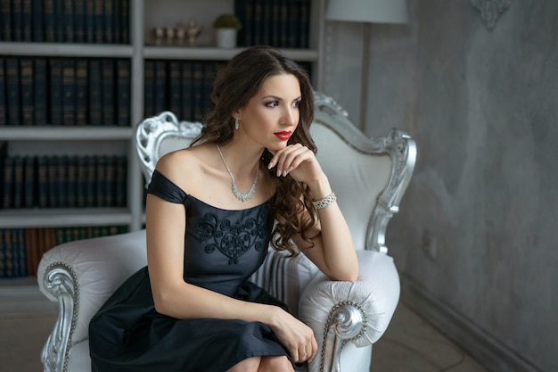 Une belle femme avec du maquillage et du rouge à lèvres est assise dans une robe noire sur une chaise blanche