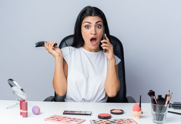 Une belle femme effrayée est assise à table avec des outils de maquillage tenant un peigne parle au téléphone