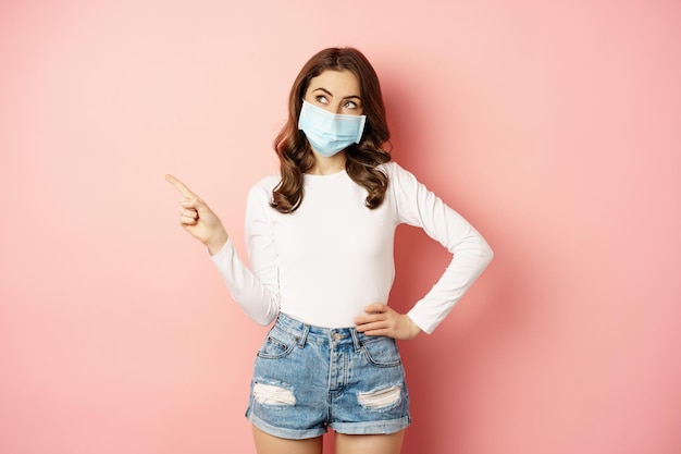 Belle femme élégante dans un masque médical de covid, pointant le doigt dans le coin supérieur gauche, montrant une publicité, debout sur fond rose