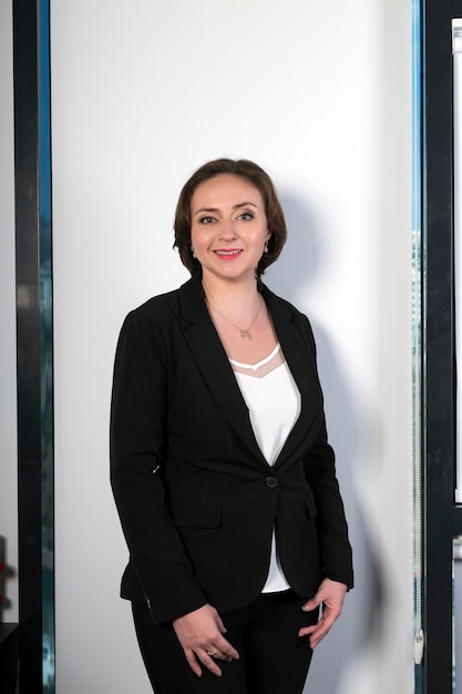 Belle femme élégante debout près de la fenêtre au bureau Adorable dame brune debout dans une veste élégante regardant la caméra avec un sourire énigmatique Belle femme professionnelle élégante en costume noir