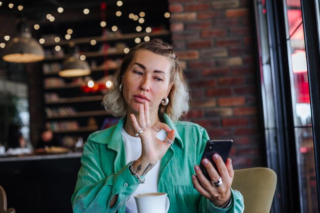 Belle femme élégante heureuse en chemise verte assise dans un café loft buvant un cappuccino faisant un appel vidéo avec son téléphone portable et montrant un signe ok