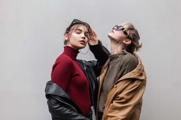 Belle femme élégante de jeunes copines dans une veste en cuir à la mode et un pull en tricot avec des lunettes de soleil se tiennent sur un fond gris dans la ville