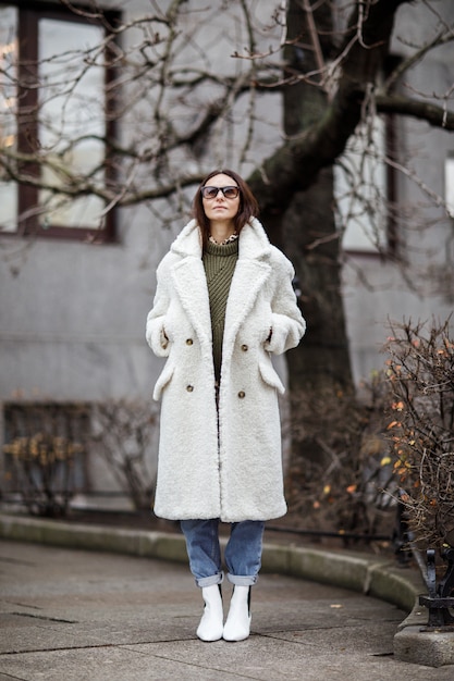 Photo belle femme élégante, marchant dans la rue par une froide journée d'hiver enneigée. fille à la mode portant un chemisier noir et un pantalon et un manteau de fourrure blanche.