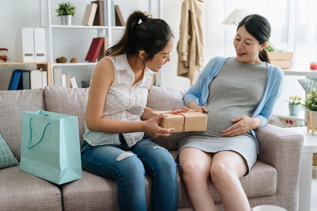 Belle femme enceinte et amie célébrant la douche de bébé dans le salon à la maison. deux jeunes femmes assises sur un canapé avec sac à provisions et boîte-cadeau. tante envoie un cadeau au gros ventre à l'intérieur de bébé.