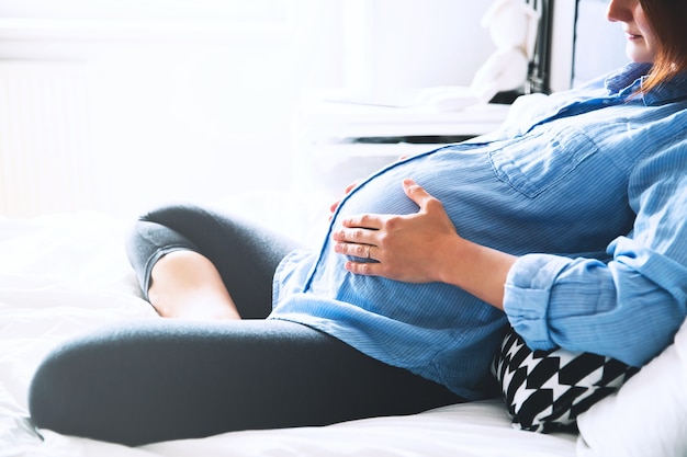 Belle femme enceinte assise au lit et tient les mains sur le ventre dans la chambre à la maison