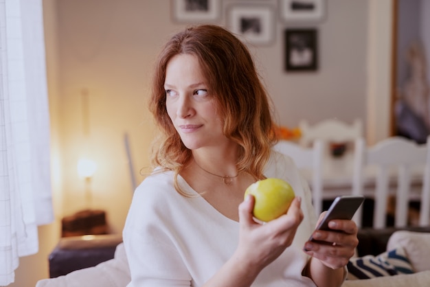 Belle femme enceinte caucasienne assise dans le salon, en utilisant un téléphone intelligent et manger des pommes.