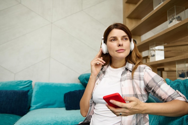 Belle femme enceinte caucasienne sereine écoutant de la musique dans des écouteurs à la maison assise sur le canapé avec un téléphone portable dans les mains et regardant rêveusement de côté Concept de grossesse et de maternité heureuse