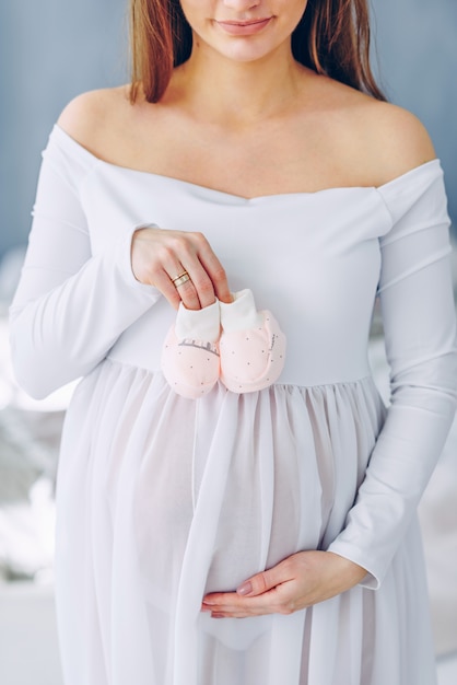 Belle femme enceinte dans une robe blanche tenant des chaussures de bébé