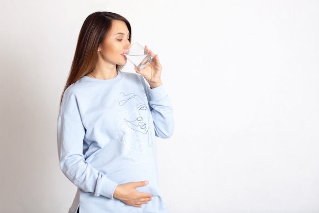 Une belle femme enceinte heureuse boit de l'eau dans un verre en verre Fond blanc