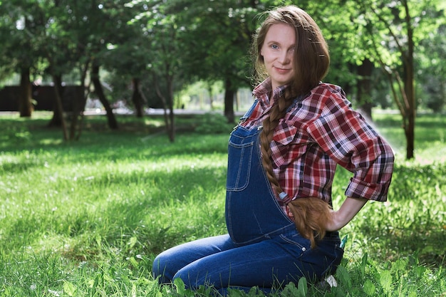 Belle femme enceinte en salopette en jean assis sur l'herbe dans le parc