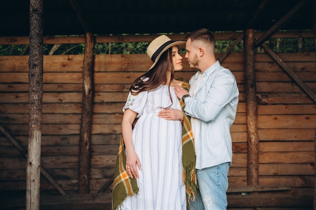 Belle femme enceinte et son mari posant près d'un bâtiment en bois. Couple heureux élégant s'aiment. Mode et style rustique.