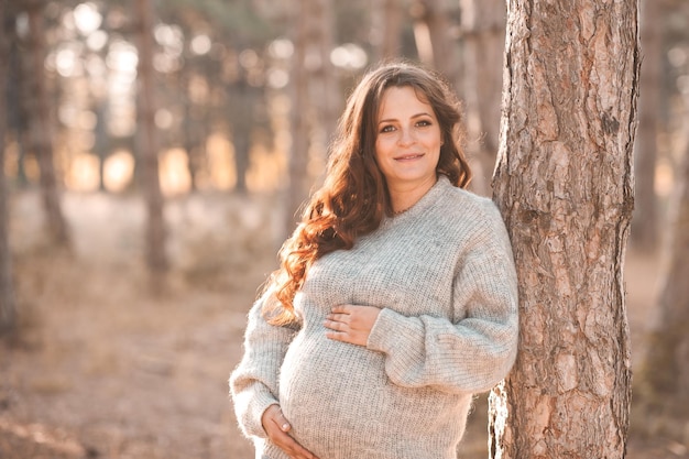 Belle femme enceinte souriante porter un cardigan en tricot posant en plein air dans les bois