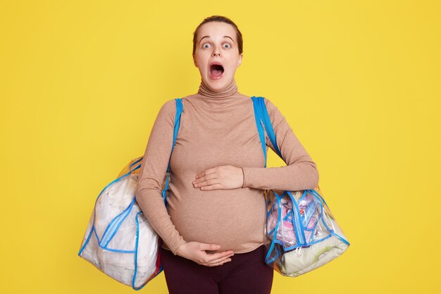 Belle femme enceinte touchant son ventre debout isolé sur un mur jaune, garde la bouche largement ouverte, ressent des contractions. doit aller à la maternité, tient des sacs avec des affaires.