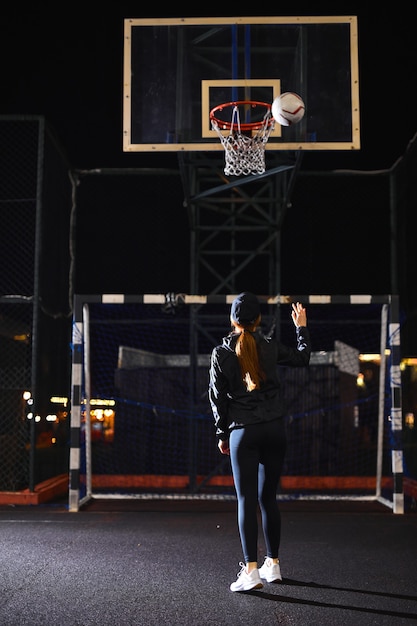 Belle femme énergique de remise en forme dans un terrain de basket-ball extérieur clôturé la nuit s'entraînant seule à lancer ...