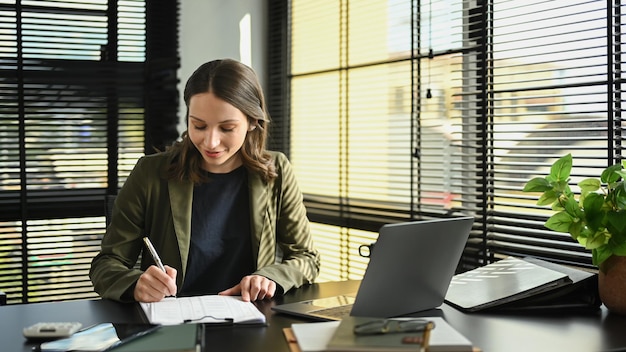 Belle femme entrepreneure caucasienne utilisant un ordinateur portable et analysant un document financier sur son lieu de travail