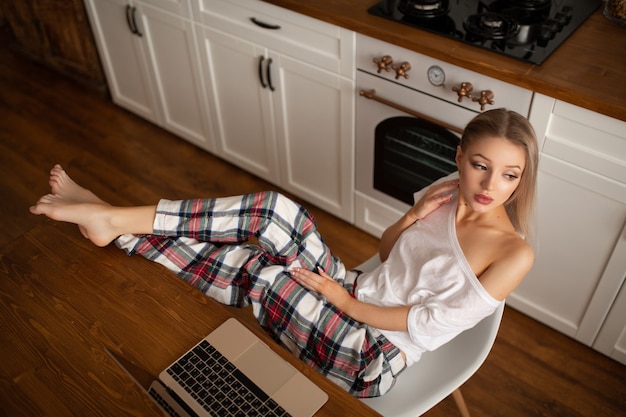 Une belle femme est assise dans la cuisine à la table à côté d'un ordinateur portable