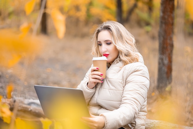 Une belle femme est assise dans un parc en automne devant un ordinateur portable et tient un verre de café à la main