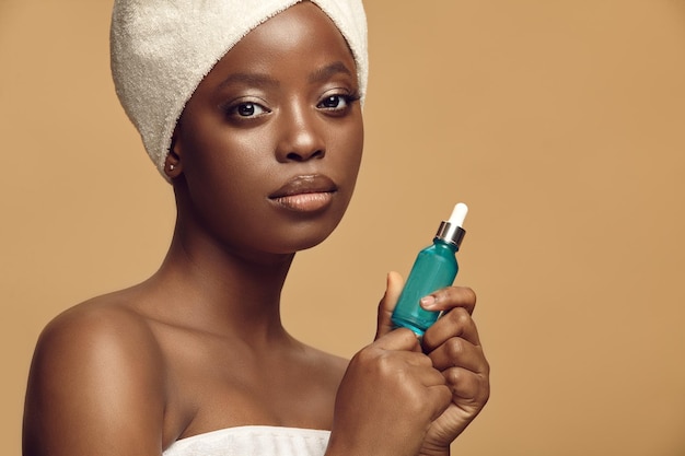 Belle femme d'ethnie afro en vêtements de spa tenant du sérum pour le visage sur fond beige