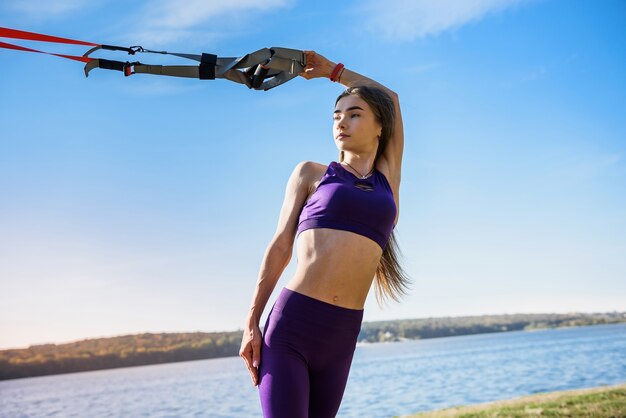 Belle femme exerçant avec des sangles de suspension TRX à l'extérieur près du lac pendant la journée. Mode de vie sain