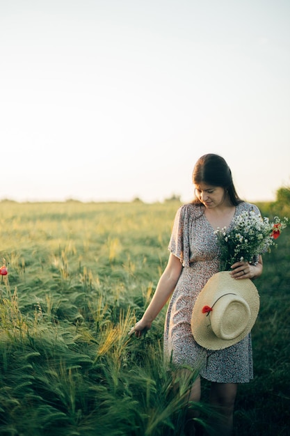 Belle femme avec des fleurs sauvages et un chapeau de paille se promenant dans un champ d'orge à la lumière du coucher du soleil Femme élégante se détendant dans la campagne d'été et rassemblant des fleurs Moment de tranquillité atmosphérique