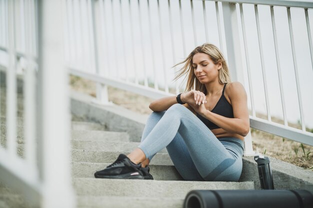 Belle femme en forme se repose avant de s'entraîner dur sur les escaliers du pont dans la ville et regarde sur smartwatch.