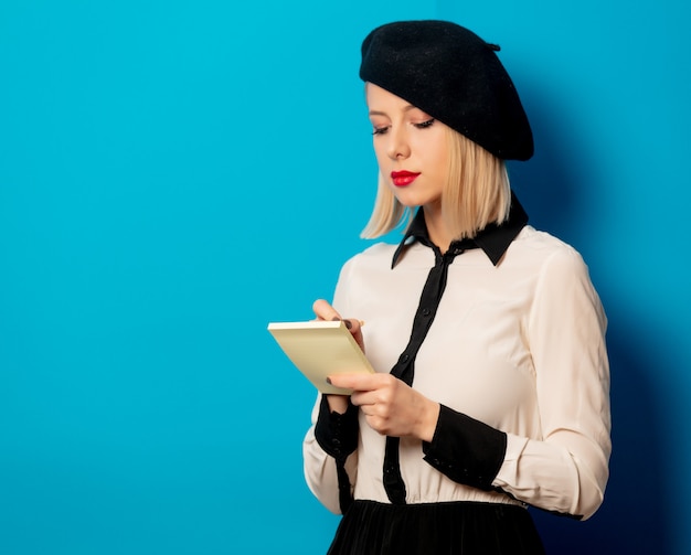 Belle femme française en béret tient un cahier avec un crayon sur le mur bleu