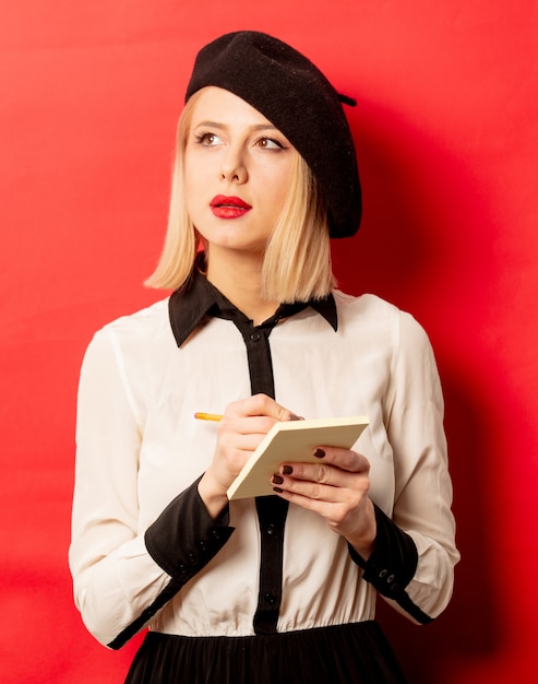 Belle femme française en béret tient un cahier avec un crayon sur un mur rouge